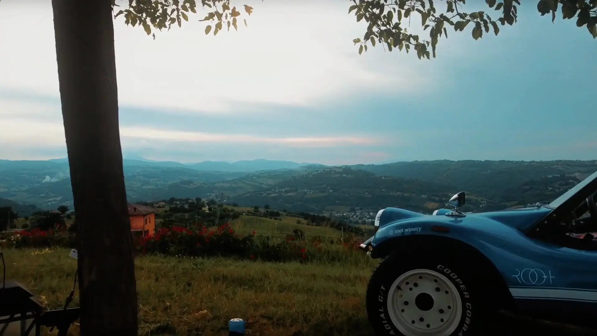 The ROOT buggy parked by a tree on the hilltop, the hills of Maiolati Spontini at dusk and a picnic table laid under the tree